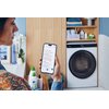Na zdjęciu widać osobę trzymającą smartfon z aplikacją wyświetlającą listę programów prania, a w tle znajduje się pralko-suszarka z widocznym logo "Candy". Obok pralki na półkach znajdują się ręczniki, butelki z detergentami oraz rośliny.
