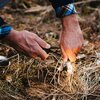 Na zdjęciu widać ręce używające krzesiwa do wytworzenia iskier nad suchą trawą na ziemi. Osoba nosi koszulę w kratę, a wokół znajduje się naturalne, leśne otoczenie.