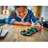 Na drewnianym stole znajduje się zielony model bolidu F1 z klocków LEGO, z widocznymi detalami aerodynamicznymi i napisami "aramco" na bokach. Obok leży ołówek i notes, a w tle widać rozmyte elementy wnętrza pomieszczenia.