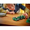 Na drewnianym stole znajduje się zielono-czarny model bolidu F1 z klocków LEGO, z widocznym napisem "KICK" na boku. Obok leży otwarty notes, a w tle widać rozmytą postać pochylającą się nad stołem.