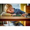Na zdjęciu znajduje się model bolidu F1 z klocków LEGO w kolorze zielonym z widocznym napisem "KICK" na boku. W tle widać osobę w niebieskiej koszuli, która patrzy na model, a na stole leżą okulary i inne przedmioty.