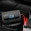 Na zdjęciu znajduje się inteligentna ładowarka z wyświetlaczem, na którym widoczne są ikony i cyfry, oraz napisy: "Qoltec SMART BATTERY CHARGER 12V 8A/24V 4A". Obok ładowarki znajdują się czerwone i czarne zaciski podłączone do akumulatora.