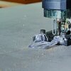 Na zdjęciu znajduje się wyrzynarka podczas pracy, tnąca powierzchnię z szarego materiału. Wokół narzędzia widoczne są drobne wióry i pył, a przezroczysta osłona zabezpiecza obszar cięcia.