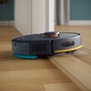 Granatowy robot sprzątający porusza się po drewnianej podłodze, pokonując niewielki próg. Na urządzeniu widoczne są elementy w kolorze pomarańczowym i niebieskim.