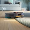Na zdjęciu znajduje się granatowy robot sprzątający na drewnianej podłodze, częściowo wjeżdżający na dywan. W tle widoczna jest szara sofa z poduszkami, a na zdjęciu umieszczono graficzne strzałki wskazujące kierunek ruchu robota.