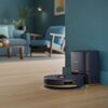 Na zdjęciu znajduje się granatowy robot sprzątający z logo "PHILIPS" na stacji dokującej, umieszczony na drewnianej podłodze w nowoczesnym wnętrzu z niebieskimi meblami. W tle widoczna jest kanapa, fotel, rośliny oraz obraz na ścianie.