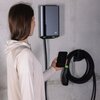 Osoba trzymająca smartfon stoi przed zamontowaną na ścianie ładowarką z logo "GC" oraz uchwytem kabla ładującego. Obok ładowarki znajduje się zwinięty kabel z zielonymi akcentami.