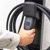 Na zdjęciu widoczny jest uchwyt kabla ładującego z logo "GC", trzymany przez rękę. Kabel jest owinięty wokół uchwytu, a tło stanowi jasna ściana.