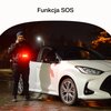 Na zdjęciu widać białe auto zaparkowane na mokrej nawierzchni nocą, obok którego stoi osoba trzymająca urządzenie emitujące czerwone światło. Na górze zdjęcia znajduje się napis "Funkcja SOS".