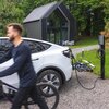 Biały samochód elektryczny jest podłączony do ładowarki Green Cell HabuDen Wallbox, stojącej na żwirowej drodze obok nowoczesnego, czarnego domu z dużymi oknami. W tle widać zielony trawnik i drzewa, a na pierwszym planie rozmytą postać na rowerze.