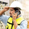 Osoba w kasku ochronnym i kamizelce odblaskowej trzyma przy uchu telefon w czarnym etui wodoodpornym TECH-PROTECT Shellbox MagSafe. W tle widoczne są rusztowania i elementy budowlane.
