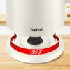 Biały czajnik elektryczny z logo "Tefal" na podstawie, która ma czerwony pasek z napisem "360°" i strzałkami wskazującymi możliwość obrotu. Tło stanowi drewniany blat i fragment kuchni.