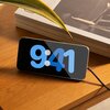 Na zdjęciu widać telefon leżący poziomo na drewnianym stole, z wyświetlaczem pokazującym godzinę 9:41 w dużych, niebieskich cyfrach. Obok telefonu znajdują się trzy książki z widocznym tytułem "CEREAL" na grzbietach, a w tle widać fragment zielonego liści
