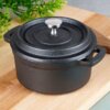 Czarny, żeliwny garnek do serwowania z pokrywką i metalowym uchwytem, umieszczony na drewnianej desce, w tle widoczne są zielone liście rośliny. Produkt ma dwa uchwyty po bokach i jest przeznaczony do serwowania potraw.