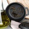 Żeliwny garnek do serwowania z pokrywką, o średnicy 10 cm i pojemności 250 ml, z widocznymi śladami oleju na dnie. Obok znajduje się szklana karafka z oliwą oraz jasna ściereczka.