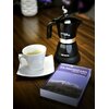 Na drewnianym stole znajduje się czarna kawiarka z napisem "Moka Aroma" i "Ariete", obok niej stoi filiżanka z kawą na białym spodku oraz książka o tytule "Kilimandżaro i Mount Meru".