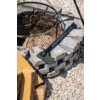 Na zdjęciu widoczna jest siekiera uniwersalna STALCO GARDEN S104210203 0.45 kg, leżąca na stosie szarych kamieni obok paleniska z metalowym rusztem. W tle znajdują się kawałki drewna oraz żwirowe podłoże.