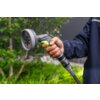 Na zdjęciu widoczny jest pistolet zraszający trzymany w dłoni, z wyraźnym napisem "STALCO GARDEN" na uchwycie. W tle znajdują się rozmyte zielone rośliny i fragmenty drzewa.