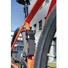 Na zdjęciu widoczne jest czarne, składane zapięcie rowerowe zamontowane na pomarańczowej ramie roweru, z widocznym mechanizmem szyfrowym. W tle znajduje się nowoczesny budynek z dużymi oknami.