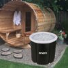 Na zdjęciu znajduje się drewniana sauna w kształcie beczki z ręcznikami wiszącymi na ścianie oraz czarna wanna do morsowania wypełniona lodem, z widocznym napisem "enero". Obok sauny znajdują się rośliny w donicach oraz kamienne ścieżki na żwirowym podłoż