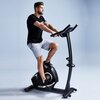 Na zdjęciu znajduje się rower elektromagnetyczny z napisem "MAXXUS" na podstawie, na którym siedzi mężczyzna w czarnej koszulce i szarych szortach. Tło jest jednolite, jasnoniebieskie.