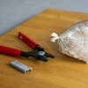 Na drewnianym blacie leży narzędzie z czerwonymi uchwytami obok siatki z zawartością, a obok znajduje się stos metalowych klipsów.