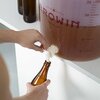 Na zdjęciu widoczny jest duży, przezroczysty pojemnik fermentacyjny z czerwonym napisem "BROWIN" oraz miarką pojemności, z którego za pomocą białego kranu nalewany jest płyn do brązowej butelki. W tle znajdują się trzy puste butelki.