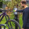 Mężczyzna trzyma zieloną kartę NFC przy stacji ładowania rowerów elektrycznych, na której widoczne jest logo "GC". Obok znajduje się rower z widoczną oponą i kierownicą, a w tle rosną drzewa.