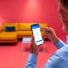 Osoba trzyma smartfon z aplikacją, na której widoczny jest napis "Smart pairing" oraz "Pairing completed" z dużym niebieskim znakiem potwierdzenia. W tle znajduje się żółta sofa z niebieską poduszką oraz stolik na różowym tle.