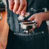 Osoba w ciemnym fartuchu trzyma portafilter z mieloną kawą, dociskając ją tamperem. W tle widoczny jest rozmyty fragment drewnianego blatu.