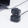 Na zdjęciu znajduje się czarna kamera internetowa z napisem "OBSBOT" na podstawie, podłączona kablem do laptopa. Kamera ma nowoczesny design z czerwonym akcentem wokół obiektywu.