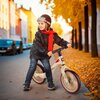 Na zdjęciu znajduje się beżowy rowerek biegowy marki NILS FUN RB138, ustawiony na tle jesiennej scenerii z opadłymi liśćmi i drzewami. Na ramie rowerka widoczny jest napis "NILS FUN".