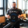 Na zdjęciu znajduje się osoba trzymająca czarną tarczę obciążeniową z niebieskim napisem "HMS 20 KG". Osoba ma na sobie czarną koszulkę z logo "HMS" i znajduje się w siłowni z widocznym sprzętem do ćwiczeń w tle.