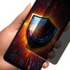 Na zdjęciu widać dłoń trzymającą smartfon z wyświetlaczem, na którym znajduje się grafika przedstawiająca oko wkomponowane w tarczę, otoczone dynamicznymi, kolorowymi wzorami. Na ekranie widoczna jest również przezroczysta folia ochronna, która częściowo 