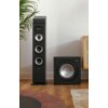 Na zdjęciu znajduje się czarny subwoofer marki Polk Audio obok kolumny głośnikowej, ustawione na drewnianej podłodze w jasnym pomieszczeniu. W tle widoczna jest część rośliny doniczkowej oraz fragment mebla z plecionką.