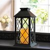 Czarna lampa ogrodowa w kształcie latarni z krzyżowym wzorem na szybach, wewnątrz której znajduje się świeca emitująca ciepłe, żółte światło. W tle widoczne są rozmyte elementy wnętrza, w tym wazon z białymi kwiatami oraz okno z widokiem na zieleń.