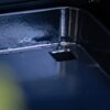 Zbliżenie na metalową powierzchnię grilla z widocznym otworem w rogu. Powierzchnia jest gładka i błyszcząca, odbijająca światło.
