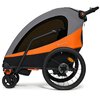Przyczepka rowerowa w kolorze pomarańczowym i szarym z czarnymi elementami, wyposażona w trzy koła, z czego dwa są większe i jedno mniejsze. Na pomarańczowej części widoczny jest napis "Kinderkraft".