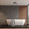 Nowoczesna łazienka z wolnostojącą białą wanną na podwyższeniu, z szarym ręcznikiem w zygzakowaty wzór, na tle ściany z szarych cegieł i drewnianych paneli. Na suficie zamontowana jest czarna lampa sufitowa z dwoma reflektorami.