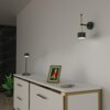 Na zdjęciu znajduje się nowoczesny kinkiet w kolorze zielono-złotym zamontowany na ścianie nad białą komodą z złotymi akcentami. Na komodzie stoi zielona lampka, ramka ze zdjęciem oraz leżą klucze, a obok znajduje się roślina w doniczce.