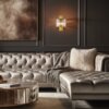 Na zdjęciu znajduje się elegancki salon z pikowaną, beżową sofą narożną, na której leży szary koc i jasna poduszka. Na ścianie wisi złoty kinkiet z przezroczystymi elementami, a obok znajduje się okrągły stolik z lustrzanym wykończeniem.