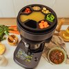 Na zdjęciu znajduje się wielofunkcyjny robot kuchenny z wyświetlaczem dotykowym, na którym widoczne są różne opcje gotowania. Na górze urządzenia znajduje się taca z podziałkami, wypełniona różnymi składnikami, takimi jak pokrojone warzywa i nasiona.