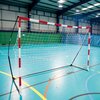 Na zdjęciu znajduje się bramka do piłki ręcznej z czerwono-białymi słupkami i siatką, ustawiona na niebieskiej podłodze hali sportowej. W tle widoczne są zielone trybuny oraz ściany hali z metalową konstrukcją.