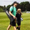 Mężczyzna w czapce i koszulce niesie torbę z napisem "QUICKPLAY" na boisku piłkarskim, obok niego idzie dziecko trzymające piłkę. W tle widoczne są drzewa i bramki do piłki nożnej.