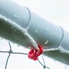 Zbliżenie na fragment białej rurki z siatką przymocowaną za pomocą czerwonego klipsa, na powierzchni rurki widoczne są krople wody. Tło jest jasne i rozmyte.