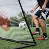 Na zdjęciu znajduje się trenażer do piłki nożnej QUICKPLAY Spot Rebounder o wymiarach 150 x 100 cm, ustawiony na zielonym boisku. Widać piłkę odbijającą się od siatki trenażera oraz fragment osoby w sportowym stroju.