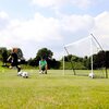 Na zdjęciu znajduje się bramka do piłki nożnej QUICKPLAY Kickster o wymiarach 300 x 200 x 100 cm ustawiona na trawiastym boisku. W tle widoczne są drzewa i niebo z chmurami.