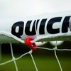 Zbliżenie na fragment białej rurki z czarnym napisem "QUICK" oraz siatkę przymocowaną czerwonym klipsem. W tle widoczna jest rozmyta zieleń.