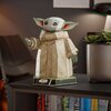 Na zdjęciu znajduje się model 3D przedstawiający postać z dużymi uszami i zieloną skórą, ubrany w beżowy strój, stojący na podstawce z napisem "Star Wars Grogu". Obok modelu znajduje się doniczka z rośliną, a w tle widoczne są książki na półce.