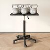 Na zdjęciu znajduje się stolik pod laptopa z czarnym blatem, na którym ustawiono kilka szarych kettlebell o wadze 10 kg z napisem "10LB 4.5KG". Stolik ma regulowaną wysokość i stoi na pięcioramiennej podstawie z kółkami, na drewnianej podłodze.